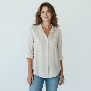 A woman with brown hair, smiling on a sunlit cobblestone street with blurred outdoor café tables in the background, wears the FRANK & EILEEN EILEEN Relaxed Button Up in Italian Cotton Tan with Textured Grid paired with jeans.