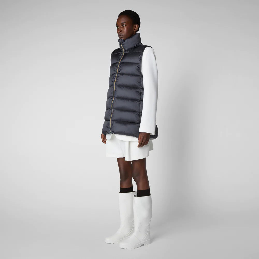 A person wearing a black cap, sunglasses, the SAVE THE DUCK - CORAL PUFFER VEST with animal-free down from SAVE THE DUCK over a black long-sleeve top, beige wide-legged pants, and black ankle boots stands against a plain background. The outfit appears to be styled for colder weather.