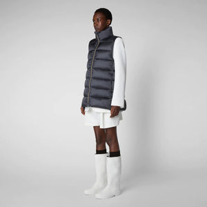 A person wearing a black cap, sunglasses, the SAVE THE DUCK - CORAL PUFFER VEST with animal-free down from SAVE THE DUCK over a black long-sleeve top, beige wide-legged pants, and black ankle boots stands against a plain background. The outfit appears to be styled for colder weather.