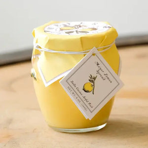 A jar of BELLA CUCINA - MEYER LEMON SPREAD adorned with bright yellow wrapping and a small tag tied around the top with string. The tag features an illustration of a lemon along with the text "Meyer Lemon spread." The jar is displayed on a reflective surface against a light background, making it perfect for enhancing any sweet tart or dessert.