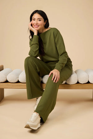 A woman wears a SOFTIES - DREAMTECH BOAT NECK WITH PINTUCK PANT SET in olive green, featuring a long-sleeve boat neck top and matching wide-leg pintuck pants.