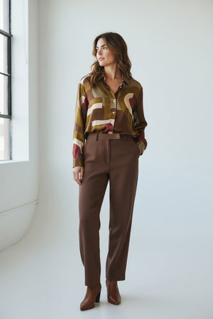 A woman with wavy brown hair wears the HARTFORD Cinema Shirt in viscose with a brown and raspberry print, looking to the side and smiling softly against a plain light background.