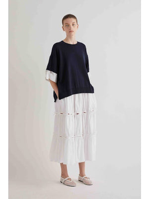 A person stands against a plain light background, wearing a loose navy top over the MEIMEIJ Cotton Poplin Skirt with Bow Detail. They have short hair, white flat shoes, and their hands are in their pockets.