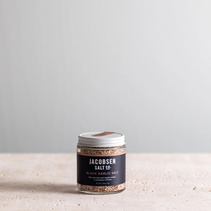 A jar of JACOBSEN SALT CO. INFUSED BLACK GARLIC SALT, featuring a black label and white lid, rests on a light beige surface against a plain light gray background.