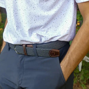 The ROOSTAS - SCOTTSDALE WOVEN STRETCH BELT by ROOSTAS is a stylish grey braided stretch belt with brown leather accents, white stitching, and a silver buckle—perfect for golf or casual wear, shown coiled on a white background.