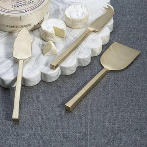 The ZODAX CHEESE SLICER AND SERVER SET includes three gold-toned cheese knives—flat spatula, chisel, and fork-tipped—each with rectangular handles, displayed side by side on a white background.
