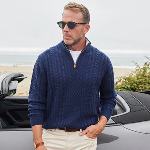 A man in sunglasses and the COLLARS AND CO - NAPOLI Cable Knit Quarter Zip Wool and Cashmere Sweater in Navy leans against a gray convertible by the beach, with the ocean and a cloudy sky behind him.