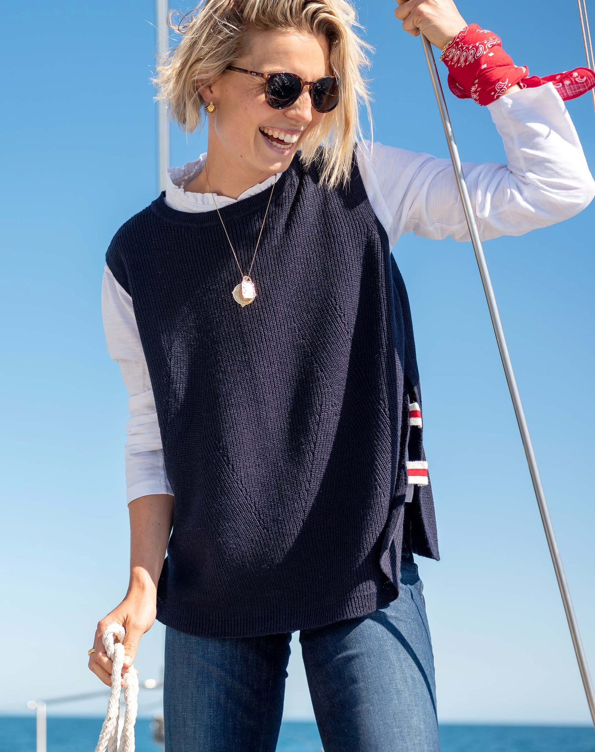 A woman stands barefoot indoors, wearing jeans, a white long-sleeve shirt, and the MERSEA Camden Vest in Navy Cherry White—an oversized sweater vest featuring a red and white striped detail down the back. She glances over her shoulder with a smile in a sunlit room.
