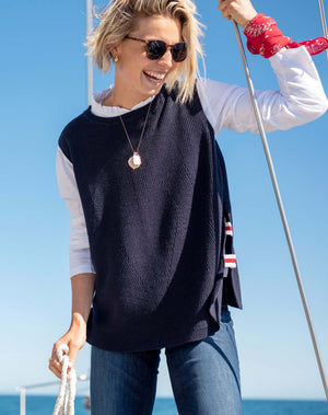 A woman stands barefoot indoors, wearing jeans, a white long-sleeve shirt, and the MERSEA Camden Vest in Navy Cherry White—an oversized sweater vest featuring a red and white striped detail down the back. She glances over her shoulder with a smile in a sunlit room.