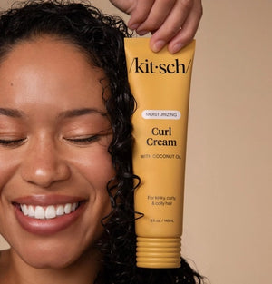 A woman with curly hair smiles and holds a yellow tube of KITSCH - MOISTURIZING CURL CREAM near her face against a neutral background, highlighting her smooth, defined curls achieved with this effective KITSCH frizz control product.