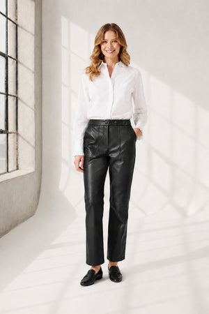 A person wearing high-waisted brown leather pants, a white long-sleeve shirt, and brown heeled ankle boots, standing against a plain light background. The person's face is not visible.