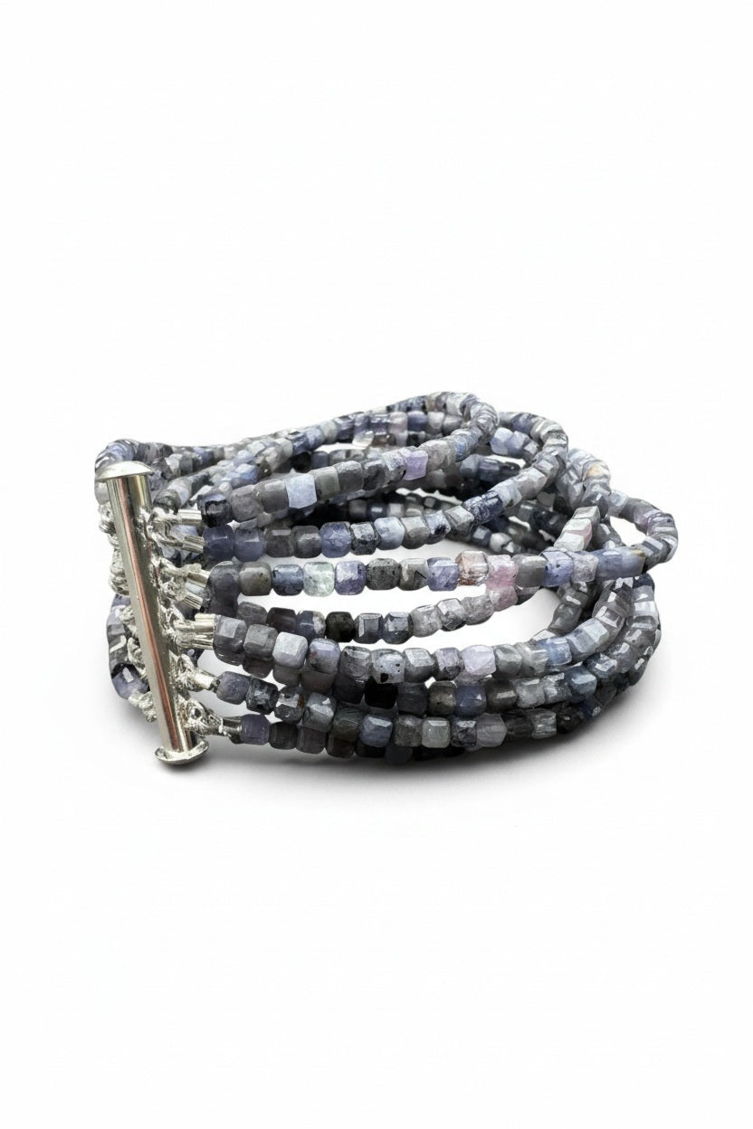 The ANN LIGHTFOOT Iolite Squares Ten Strand Bracelet features multiple coiled strands of small, faceted square-cut iolite and gray beads with a silver slide clasp, shown against a white background.