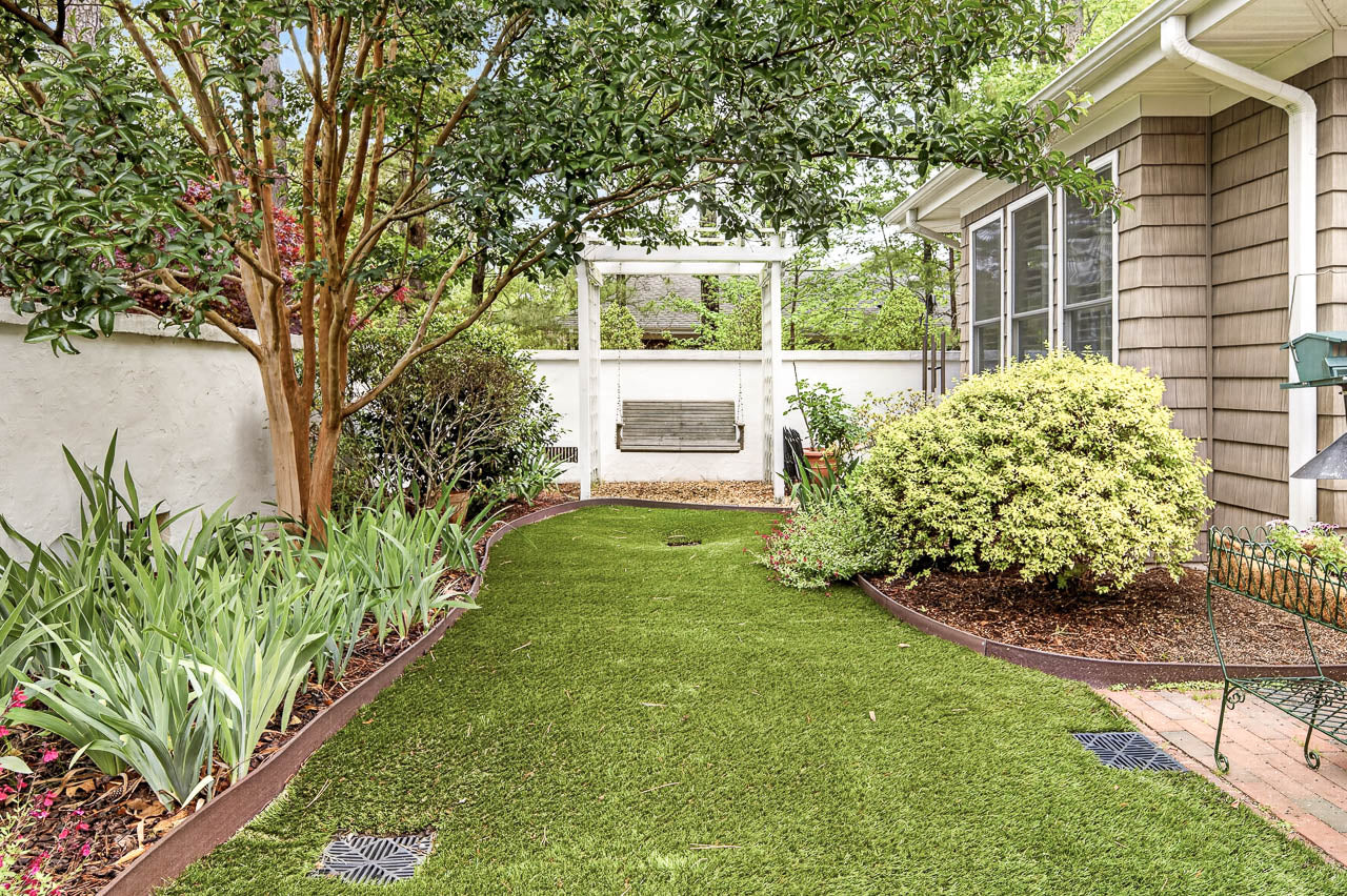 A small backyard garden with green artificial grass, flowering plants, shrubs, and a wooden swing hanging from a white arbor near a tan house with large windows.