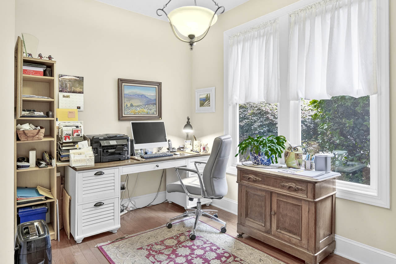 Bright home office with a white desk, gray chair, printer, and computer. Shelves hold books and supplies. Large windows with white curtains let in natural light. A wooden cabinet and a potted plant sit by the window.