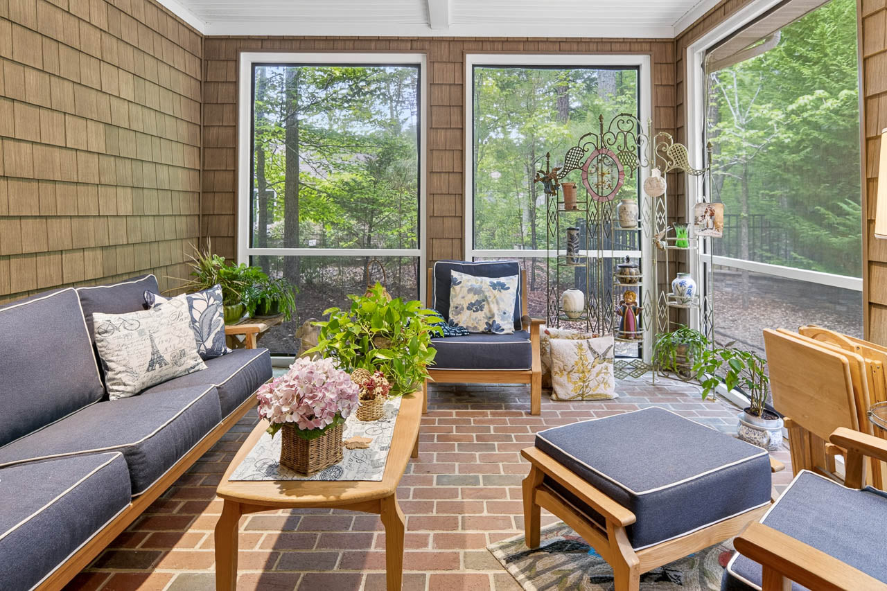 A cozy screened-in porch with cushioned wooden furniture, potted plants, a coffee table with flowers, and views of green trees outside. The floor is brick, and decorative items are displayed on metal shelves.