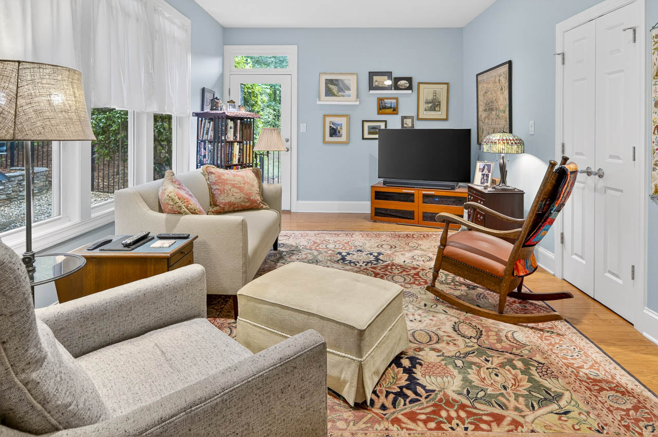 A cozy living room with two armchairs, an ottoman, a wooden rocking chair, a TV on a wooden stand, bookshelves, large windows with white curtains, and framed art on pale blue walls above a patterned rug.