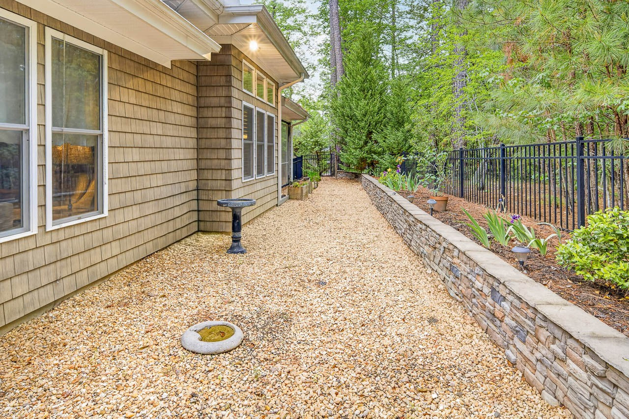 A side yard with tan shingle siding, large windows, and a gravel pathway bordered by a stone retaining wall with plants. There's a bird bath and a black metal fence along the edge, with trees in the background.