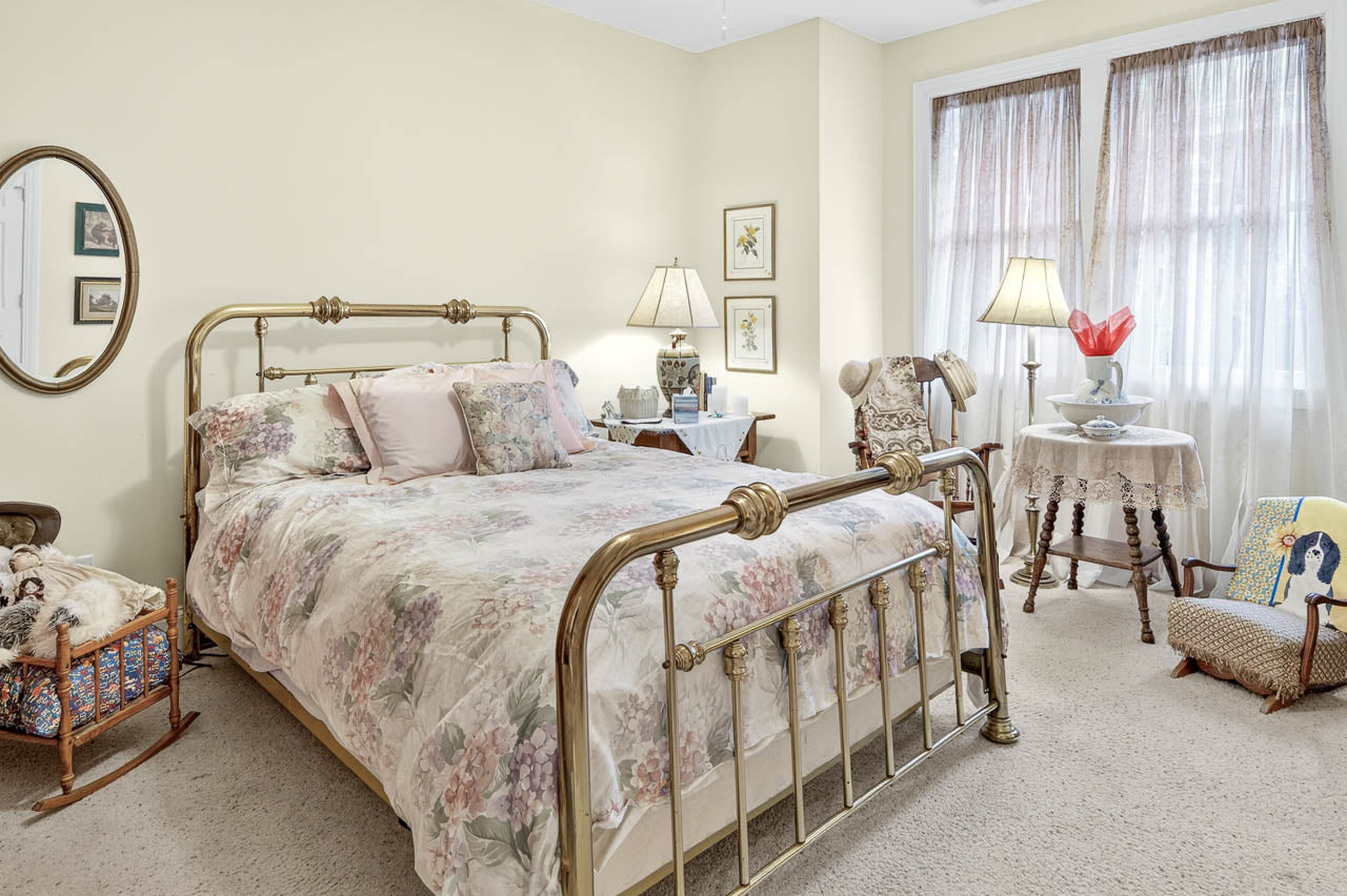 A cozy bedroom with a brass bed frame, floral bedding, vintage decor, a rocking cradle, side tables with lamps, framed art on cream walls, and soft light filtering through sheer curtains.