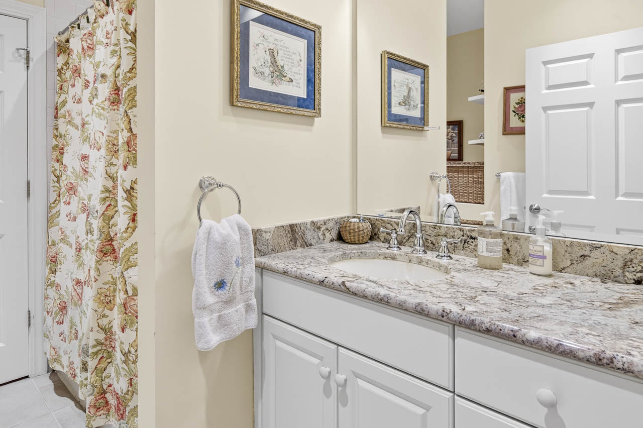 A bathroom with a granite countertop, white cabinets, a wall mirror, two framed pictures, a floral shower curtain, and a white towel hanging on a ring. Soap and lotion bottles sit near the sink.