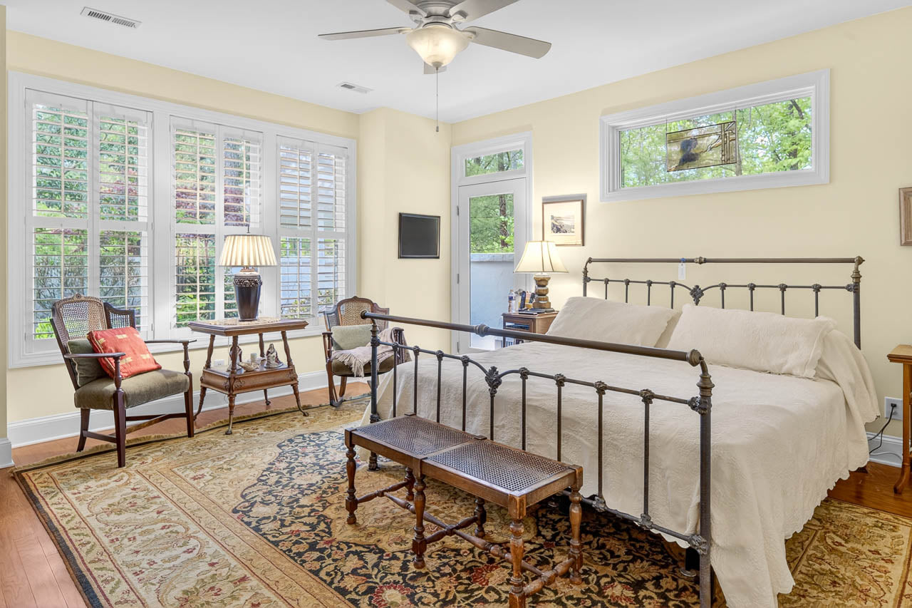 A bright bedroom with a black metal bed, neutral bedding, a wooden bench, vintage chairs, side tables with lamps, large windows with shutters, yellow walls, and a patterned rug on a hardwood floor.