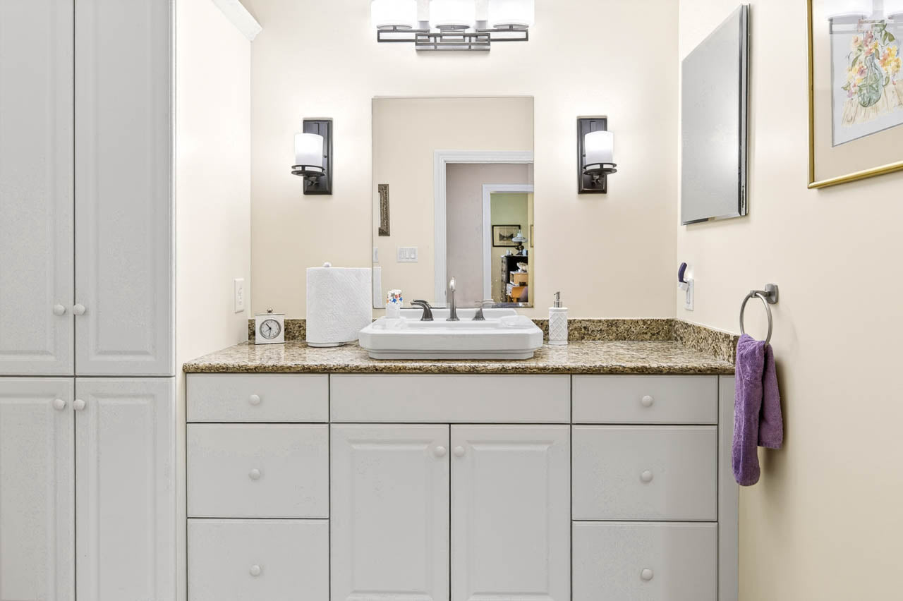 Bathroom vanity with white cabinets, granite countertop, and dual sinks. A mirror, two wall sconces, and a purple towel on a ring are visible, along with a tissue box, soap dispenser, and wall art.