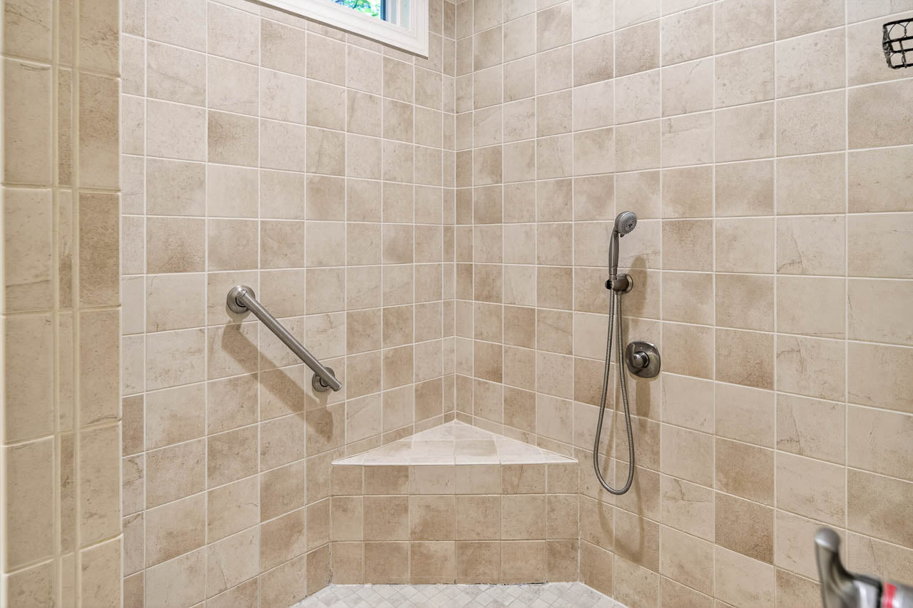 A tiled walk-in shower with a built-in corner bench, a grab bar on the left wall, a handheld showerhead, and a small window at the top left. The tiles are beige and arranged in a grid pattern.