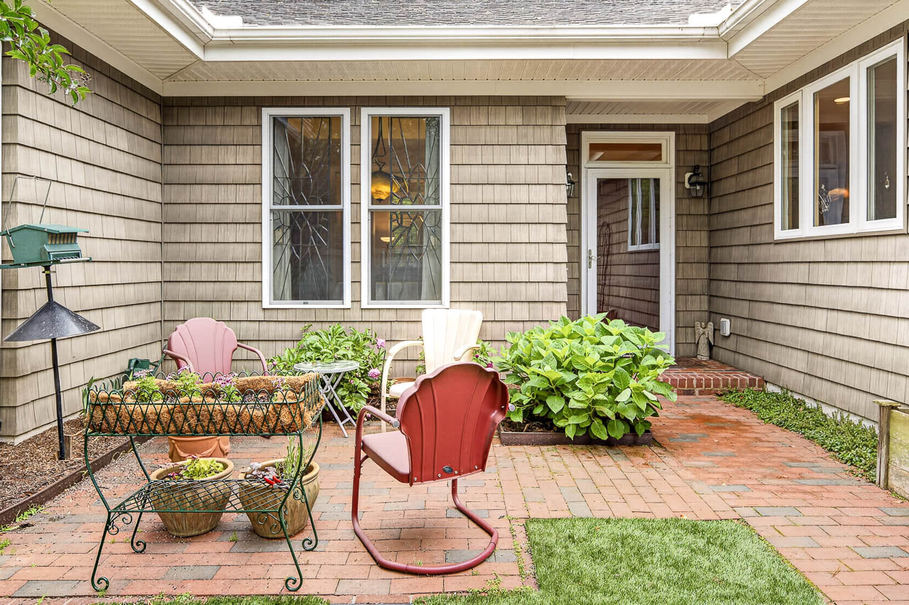 A cozy patio with three colorful chairs, a small green table with potted plants, and a brick walkway, surrounded by beige siding, windows, and greenery on a sunny day.