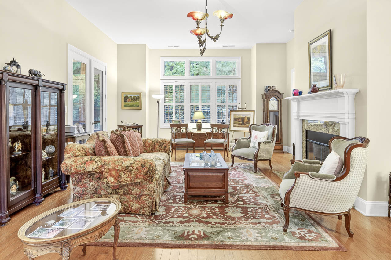 Bright, elegant living room with large windows, floral sofa, patterned rug, two armchairs, wooden coffee tables, glass display cabinet, grandfather clock, and a white fireplace with art and decor on the mantel.
