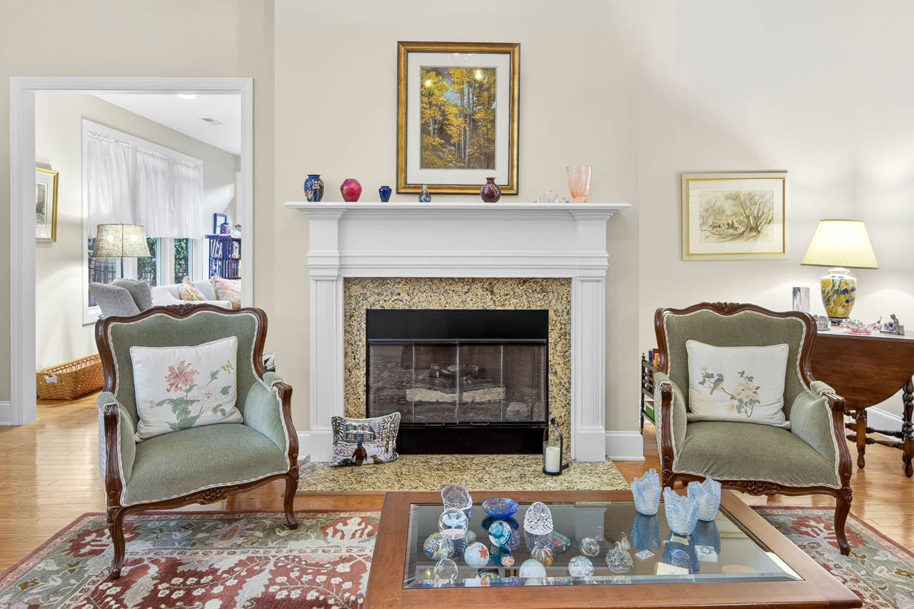 A cozy living room features two green armchairs with floral pillows facing a fireplace with a white mantle. A glass coffee table with decorative items sits on a patterned rug. Art and lamps decorate the room.