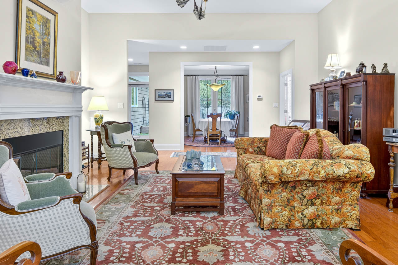 A cozy living room with a floral sofa, two armchairs, a patterned rug, a glass coffee table, a fireplace, antique cabinets, and a view into a dining area with a chandelier and wooden furniture.
