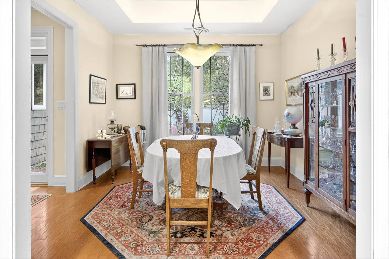 A bright dining room with a round table covered by a white cloth, six wooden chairs, a patterned rug, a glass-fronted cabinet, houseplants, and large windows with curtains letting in natural light.
