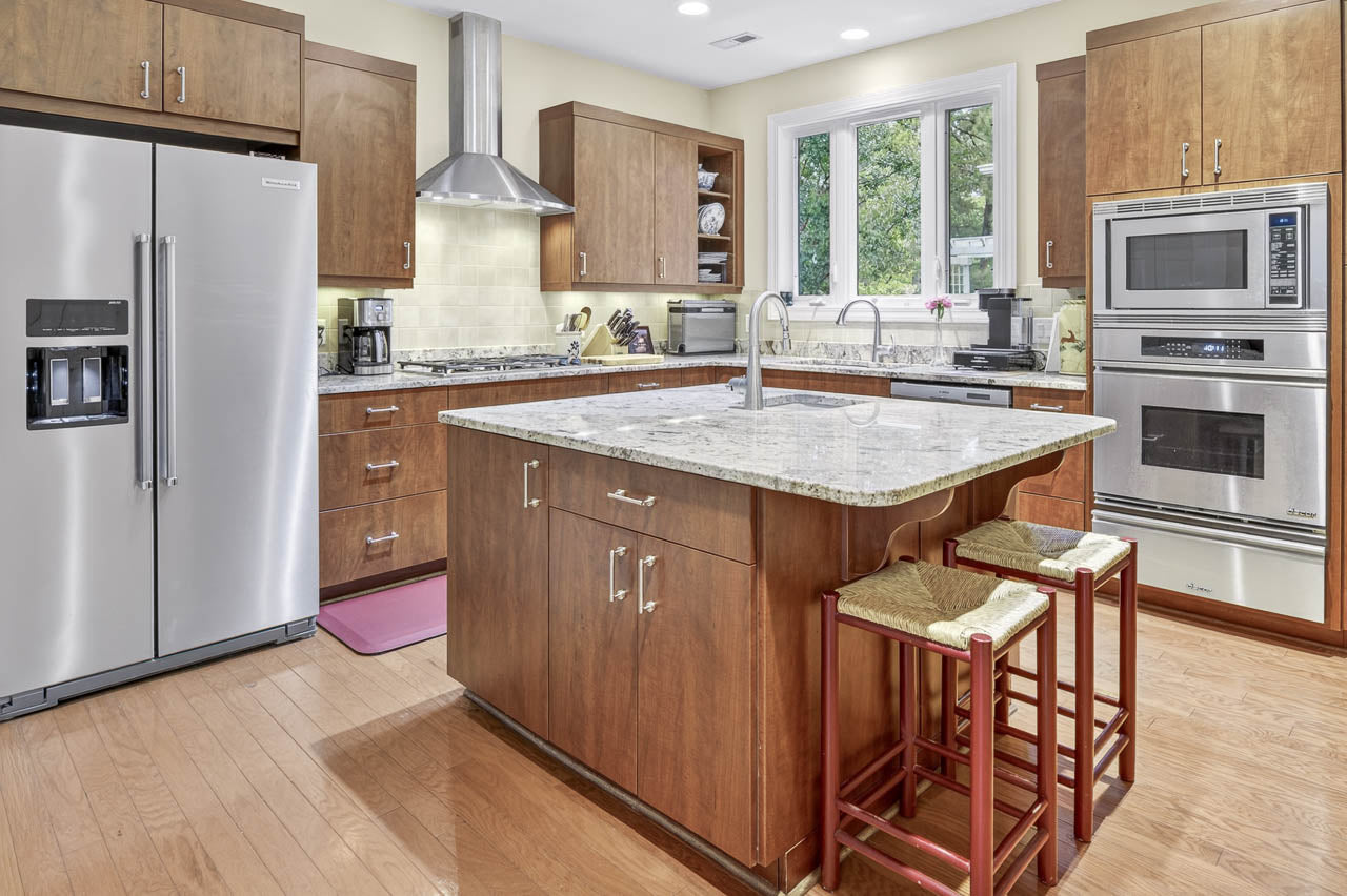 Modern kitchen with wooden cabinets, stainless steel appliances, a large island with a marble countertop, two stools, and a window above the sink letting in natural light.