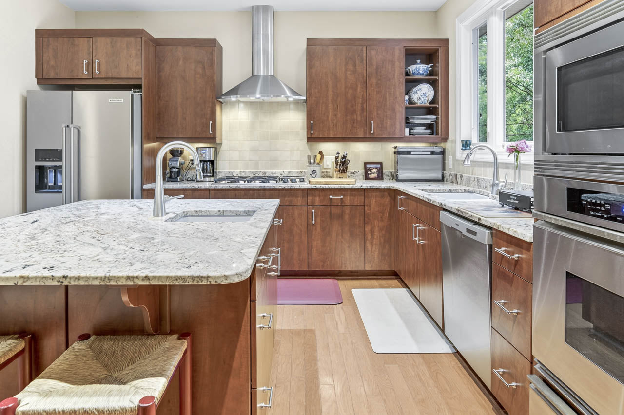 Modern kitchen with wooden cabinets, stainless steel appliances, marble countertops, a double sink island, and large window. Two stools are tucked under the island, and small kitchen appliances sit on the counter.