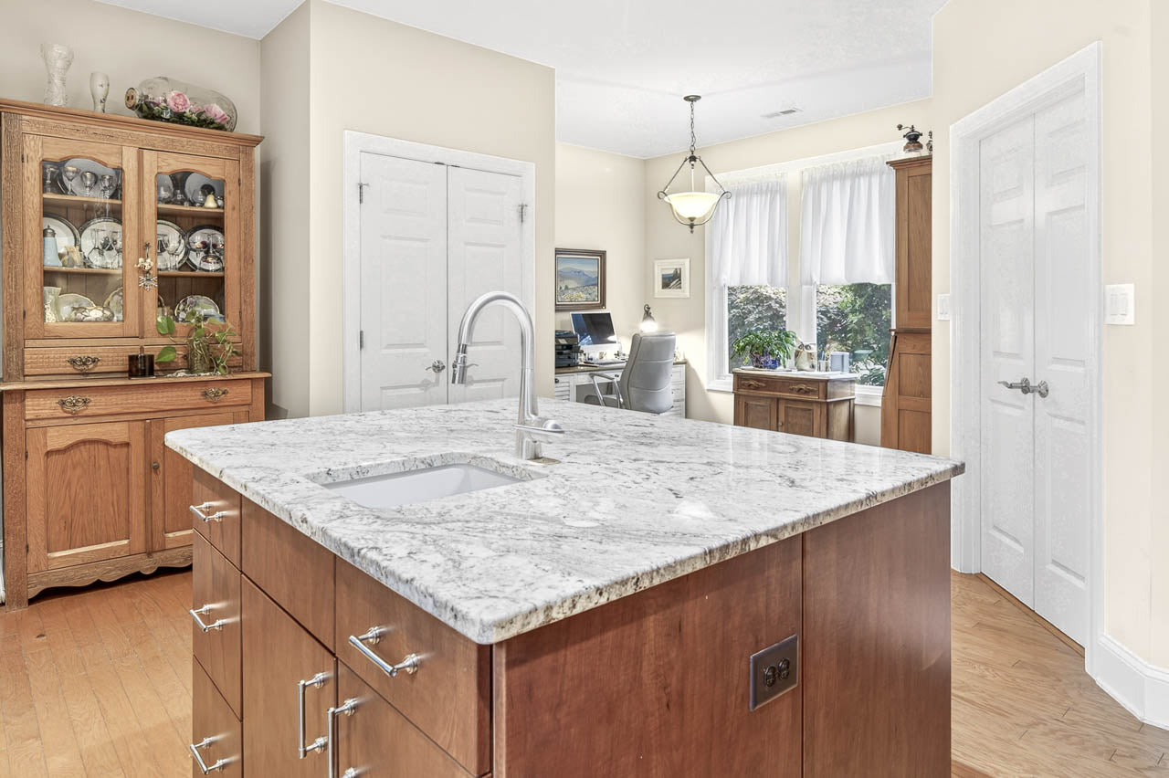 A bright kitchen with a marble island in the foreground, wooden cabinets and hutch along the walls, and a small workspace with a desk and chair by a window in the background.