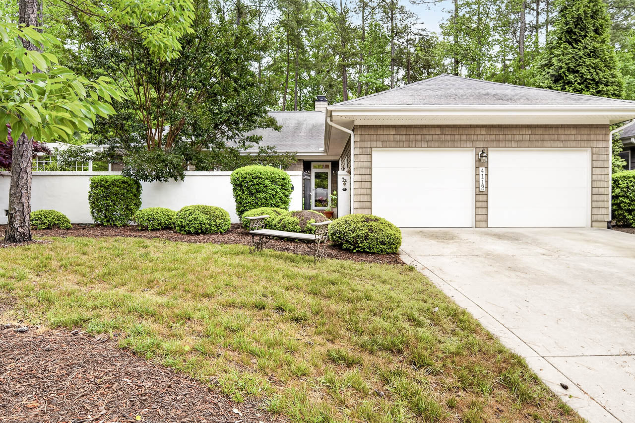 Single-story house with a double garage, light-colored siding, a gray roof, and a concrete driveway. There is green landscaping with bushes, trees, and a small bench near the entrance path.