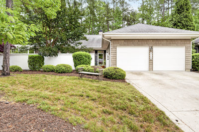 Single-story house with a double garage, light-colored siding, a gray roof, and a concrete driveway. There is green landscaping with bushes, trees, and a small bench near the entrance path.
