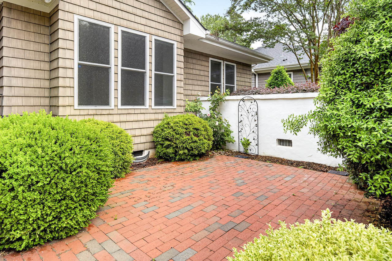 A small brick patio bordered by green bushes, next to a beige house with three large windows and a white garden wall with a decorative iron gate.