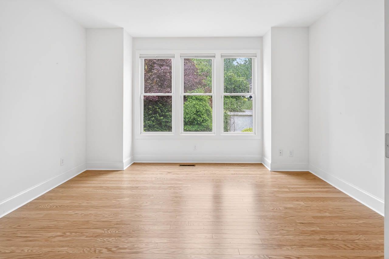 Empty room with white walls, light wood flooring, and three large windows letting in natural light. Lush green trees and shrubs are visible outside through the windows.