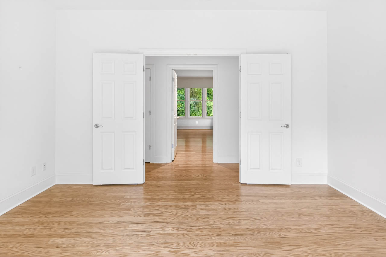 Bright, empty room with light wood floors, white walls, and double white doors open to another sunlit room with large windows and a view of green trees outside.