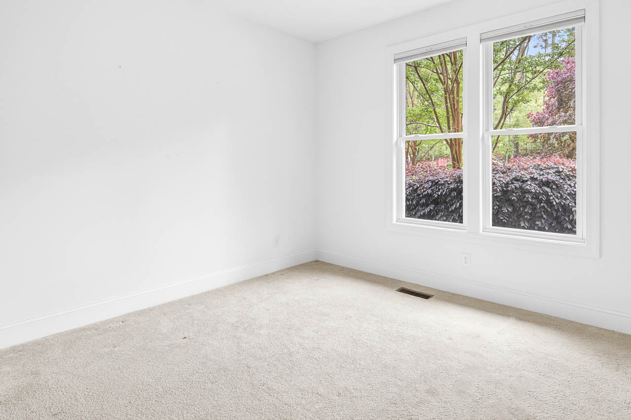 A bright, empty room with white walls and light carpeted floor. Two large windows show green trees and colorful shrubs outside, letting in natural light.