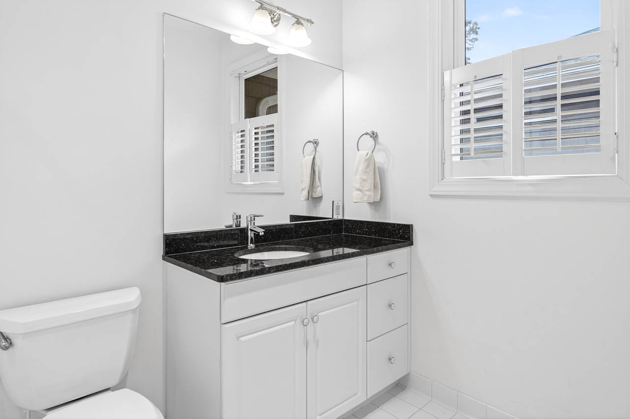 A bright, white bathroom with a black granite countertop, white vanity cabinets, a large wall mirror, a toilet, and a window with white shutters. Two towels hang on a ring holder near the sink.