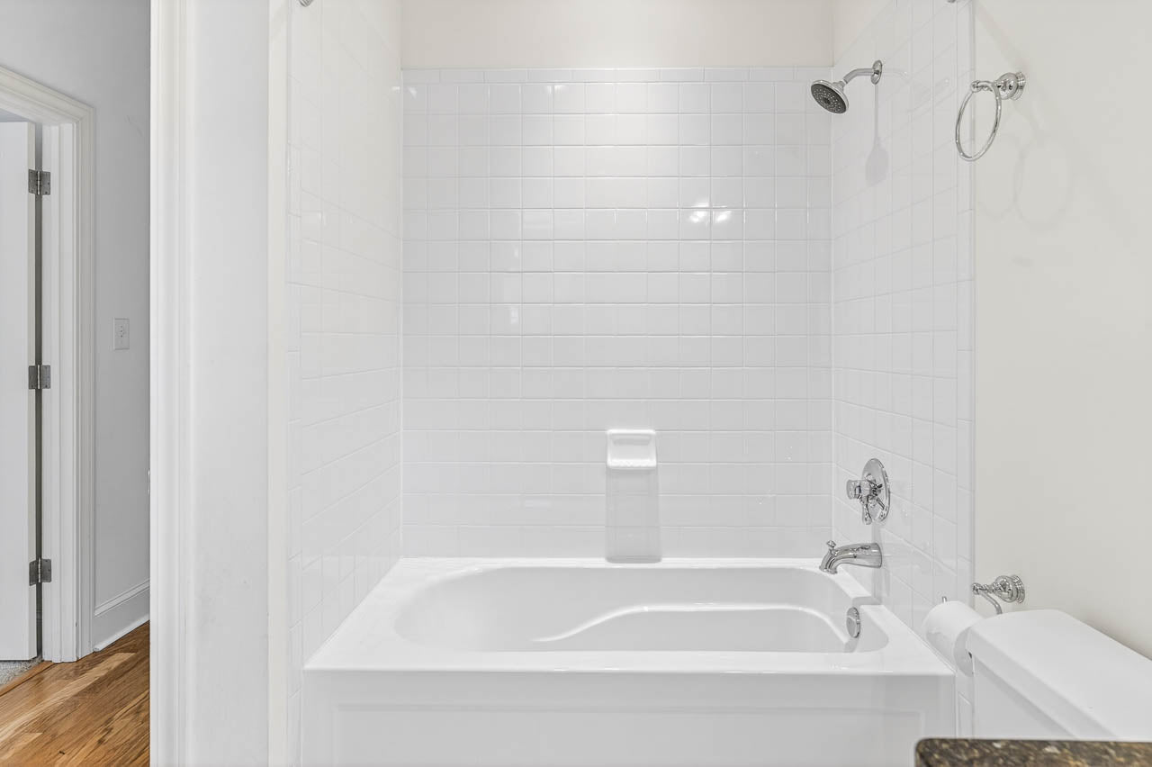 White bathroom with a built-in bathtub, white tiled walls, silver showerhead and faucet, a toilet paper holder on the back wall, and a partially visible toilet in the foreground.