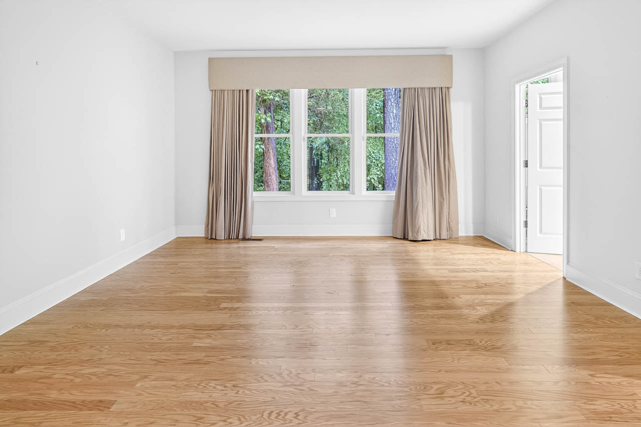 Empty room with light wood flooring, white walls, large windows with beige curtains, and a door open to the outside. Trees and greenery are visible through the windows.
