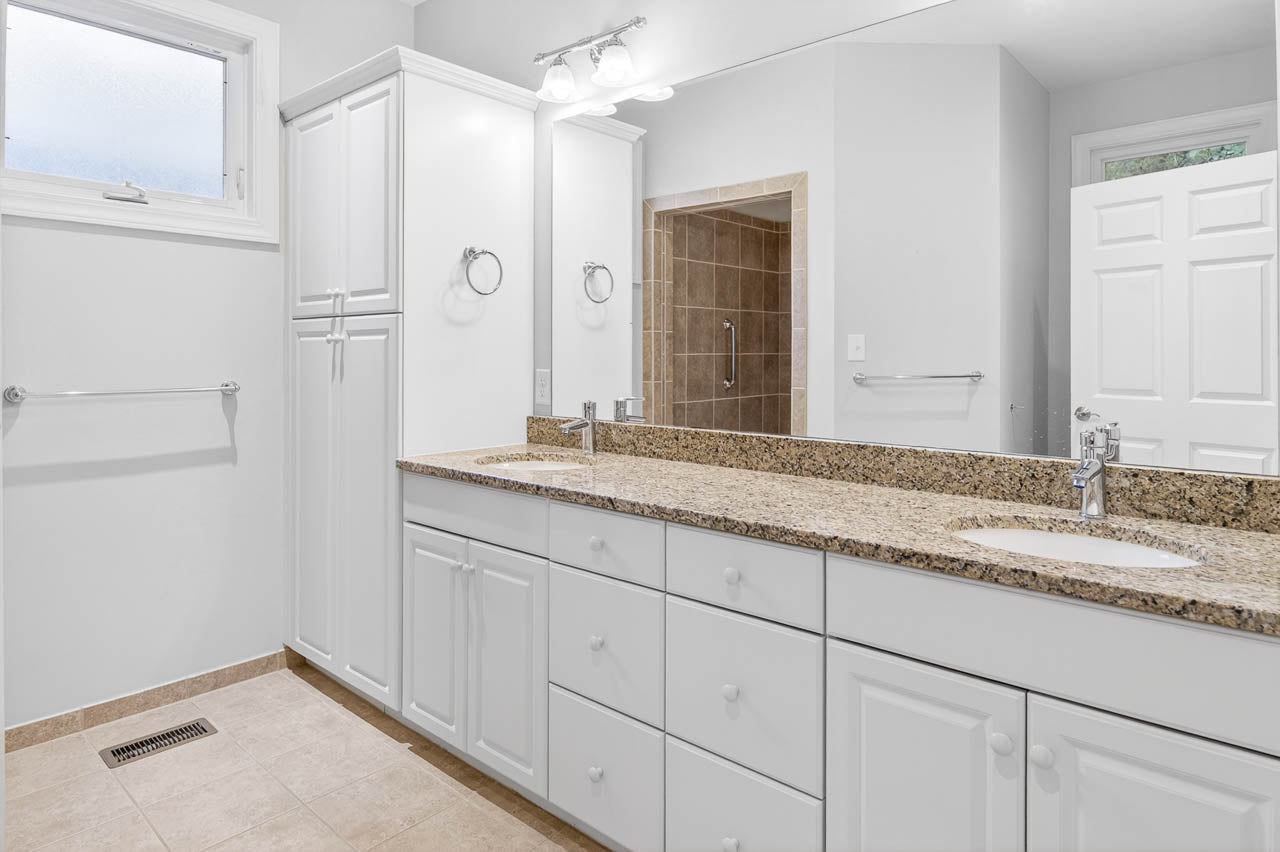 Bright, modern bathroom with white cabinets, granite countertop, double sinks with mirrors, chrome fixtures, tan tile floor, and a glass-enclosed shower area in the background.