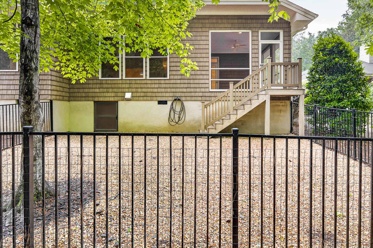 A fenced backyard with gravel ground, a tree on the left, and a house with brown siding, multiple windows, a garden hose on the wall, and stairs leading to a door.