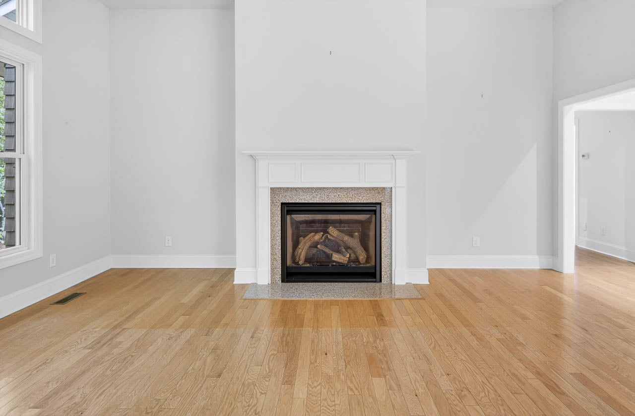 A bright, empty room with light wood floors, white walls, and a white mantel fireplace centered on the far wall. Large windows are on the left, and an open doorway leads to another space on the right.