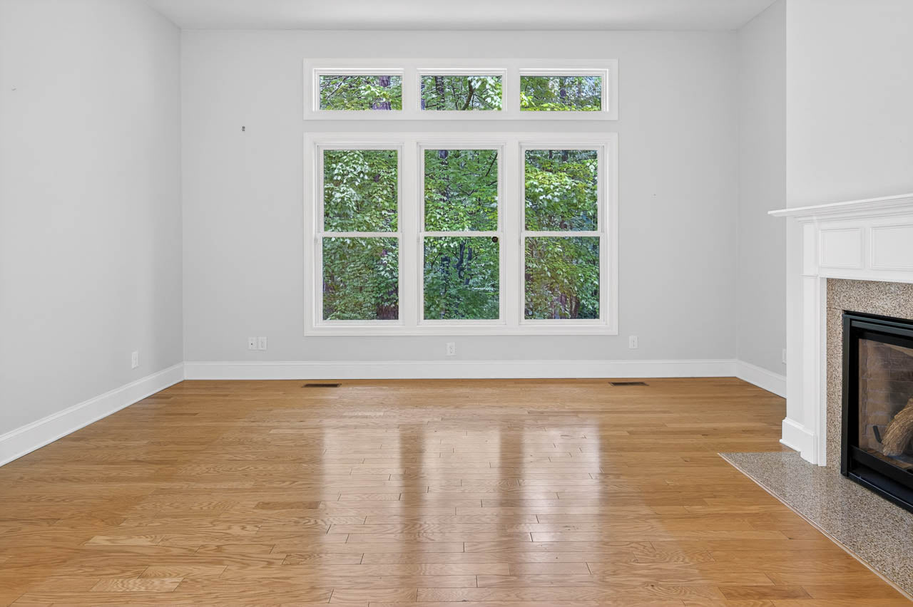 An empty living room with light hardwood floors, a white fireplace on the right, light gray walls, and large windows showing green trees outside.