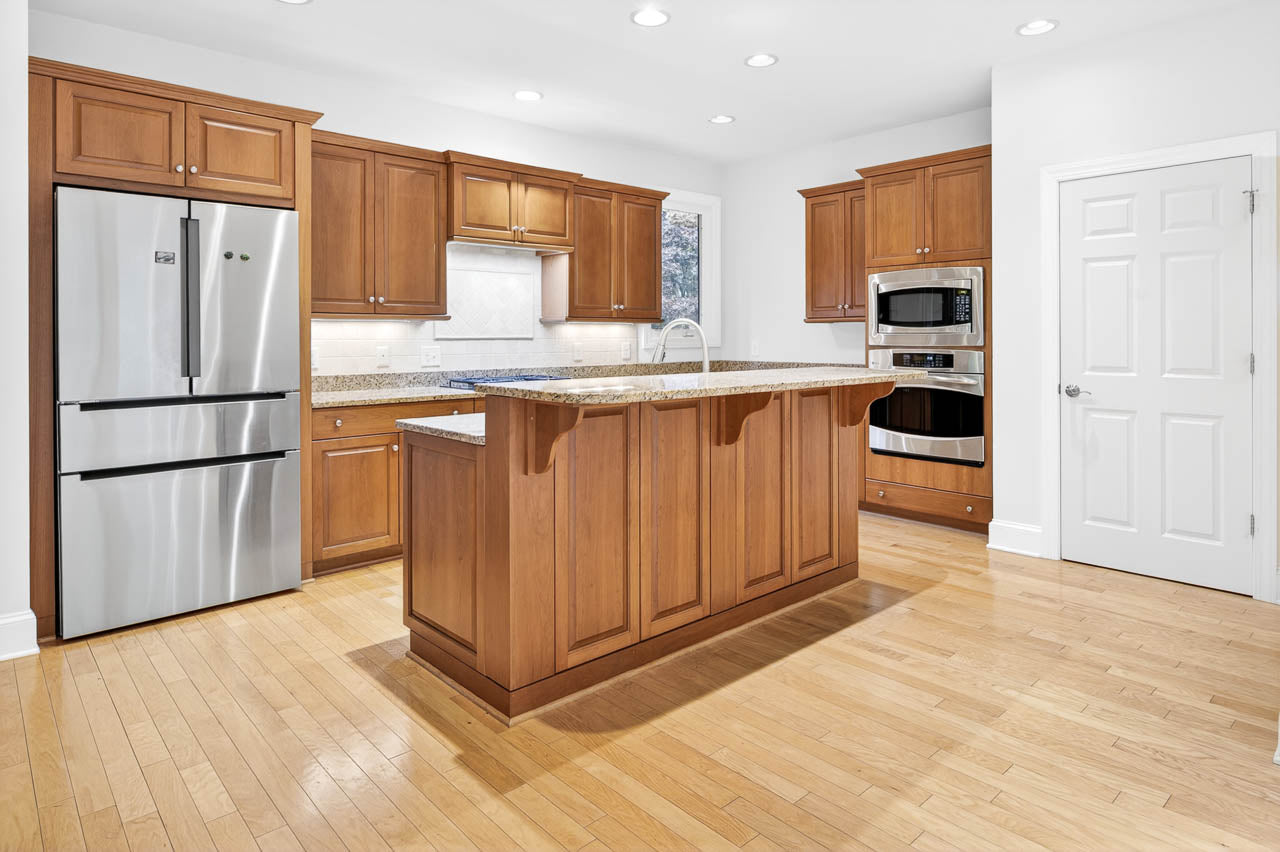 Modern kitchen with wooden cabinets, stainless steel appliances, granite countertops, a central island with an overhang, white walls, and light wood flooring. A closed white door is visible on the right side.