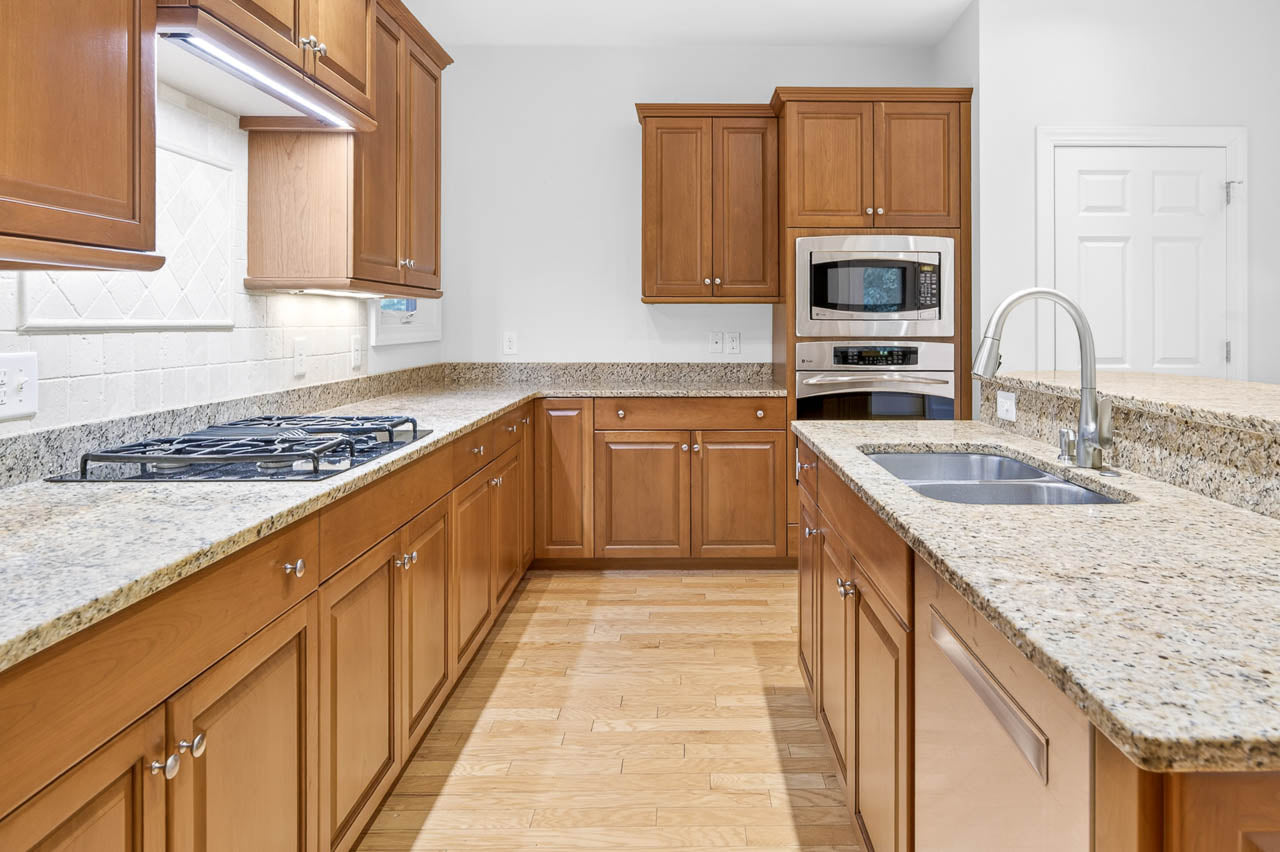 Modern kitchen with wooden cabinets, granite countertops, a gas cooktop, built-in microwave and oven, double sink with a gooseneck faucet, and light wood flooring. Walls and backsplash are white.