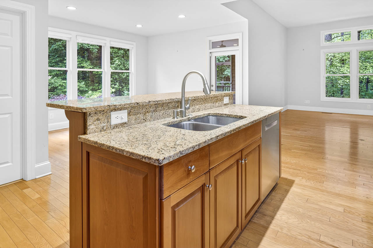 A modern kitchen island with a granite countertop, stainless steel sink, and wood cabinets, set in an open and bright room with large windows and light hardwood floors.